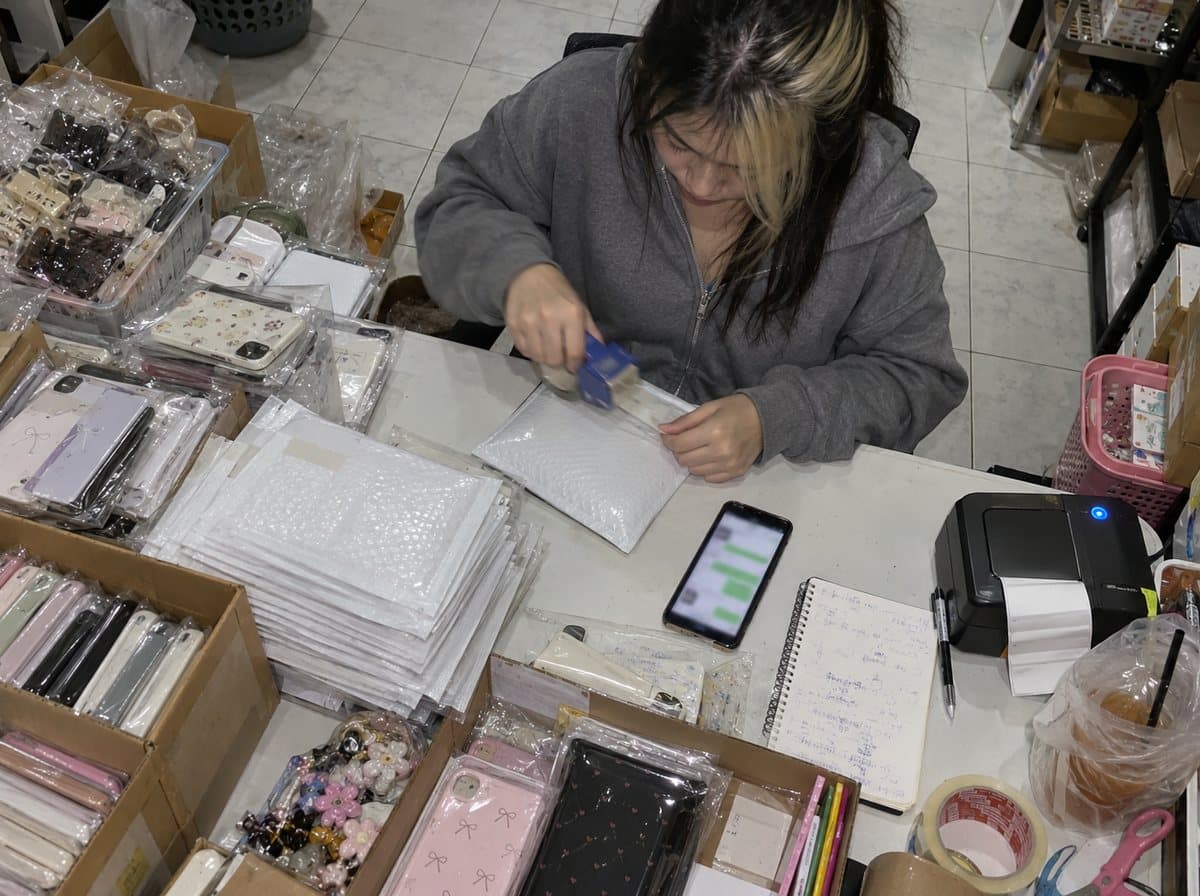 Pim, a young Thai online seller, taping a parcel shut beside boxes of phone cases and hair clips